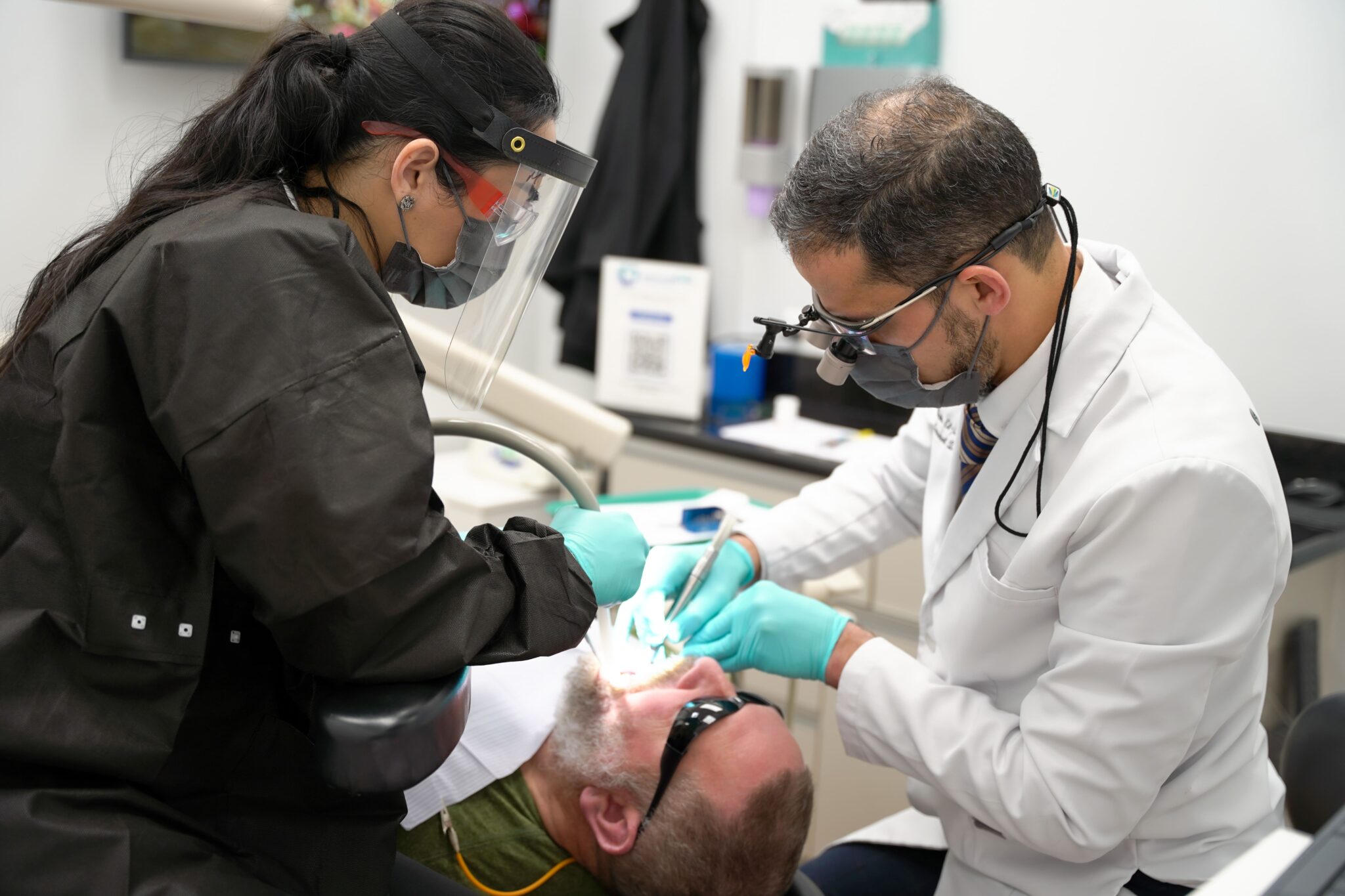 A dentist in a white lab coat uses dental tools to work on a patient lying in a dental chair, while an assistant wearing a black outfit and a face shield supports the procedure, all set in a bright dental office.
