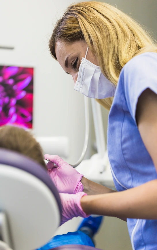A dental professional wearing a mask and pink gloves is focused on a patient's mouth during a procedure while a colorful monitor displays floral graphics in the background.
