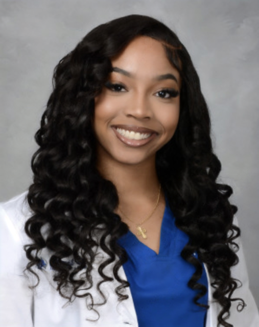 Smiling young woman with long, dark curly hair wearing a blue blouse and white coat, posing for a professional portrait.